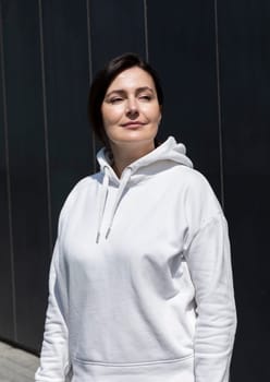 Portrait Smiling Mature Brunette Woman in White Hoodie Looking Away, Dark Background Wall. Sportive Relaxed 40 Yo Beautiful Caucasian Woman in City. Vertical Plane. High quality photo