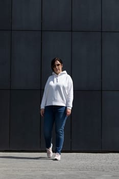 Full Length Pretty Mature Brunette Woman in White Hoodie, Denim and Sunglasses Posing On Dark Background Wall. Sportive Confident 40 Yo Beautiful Caucasian Woman in Urban Area Vertical Plane.