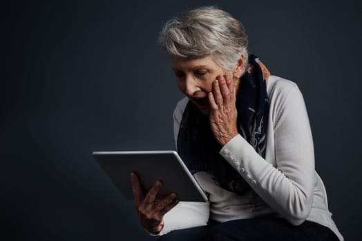 This contraption is wonderful. Studio shot of an cheerful elderly woman standing and using a tablet