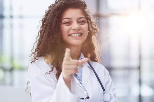 Smiling young female doctor in white coat standing and pointing forward with finger