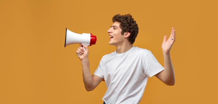 Young man shouting into megaphone making announcement against yellow background in studio
