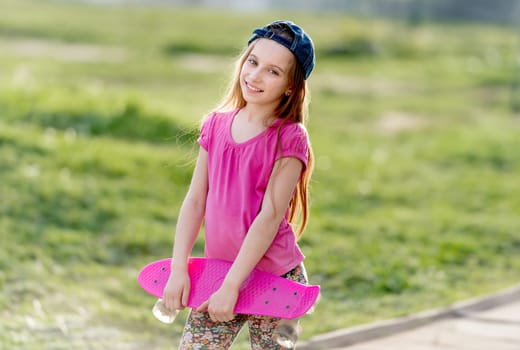 Attractive teenage girl with a cap on holding her bright pink skating board, smiling
