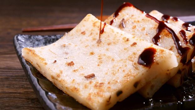 Pouring black soy sauce on ready-to-eat delicious turnip cake, Chinese traditional local dish radish cake in restaurant, close up, copy space.