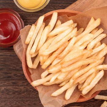 Golden yummy deep French fries on kraft baking sheet paper and serving tray to eat with ketchup and yellow mustard, top view, lifestyle.