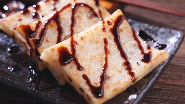 Pouring black soy sauce on ready-to-eat delicious turnip cake, Chinese traditional local dish radish cake in restaurant, close up, copy space.