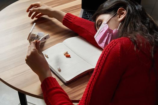 sick young woman with pills and thermometer on a table working on a laptop from home on quarantine isolation. Online work, stay home concept