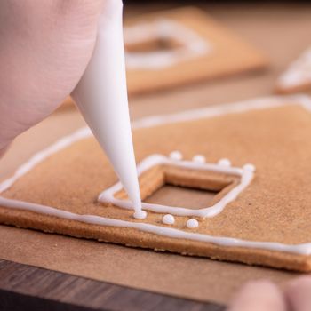 Woman is decorating gingerbread cookies house with white frosting icing cream topping on wooden table background, baking paper in kitchen, close up, macro.