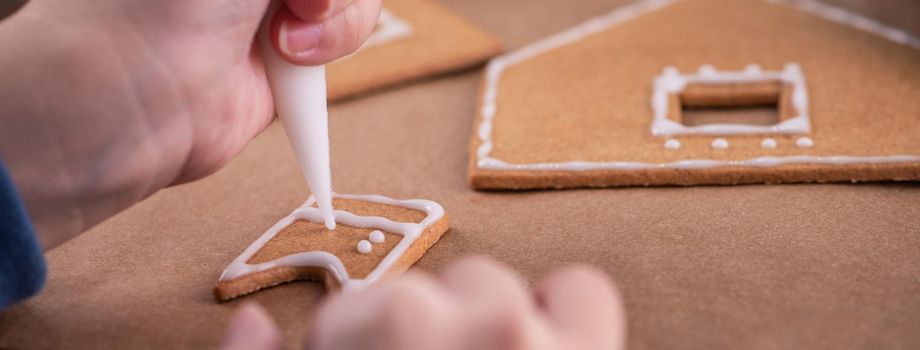 Woman is decorating gingerbread cookies house with white frosting icing cream topping on wooden table background, baking paper in kitchen, close up, macro.