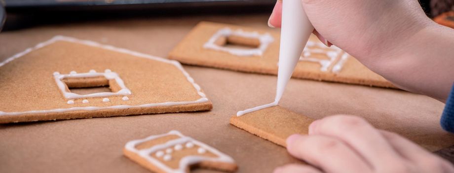 Woman is decorating gingerbread cookies house with white frosting icing cream topping on wooden table background, baking paper in kitchen, close up, macro.