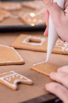 Woman is decorating gingerbread cookies house with white frosting icing cream topping on wooden table background, baking paper in kitchen, close up, macro.