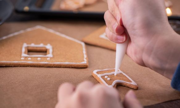 Woman is decorating gingerbread cookies house with white frosting icing cream topping on wooden table background, baking paper in kitchen, close up, macro.
