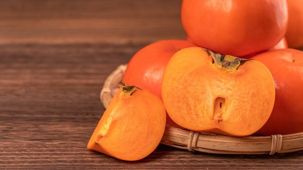 Sliced sweet persimmon kaki in a bamboo sieve basket on dark wooden table with red brick wall background, Chinese lunar new year fruit design concept, close up.