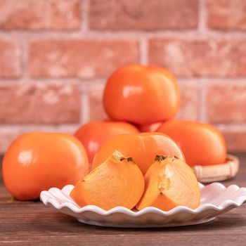 Sliced sweet persimmon kaki in a bamboo sieve basket on dark wooden table with red brick wall background, Chinese lunar new year fruit design concept, close up.