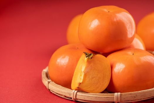 Fresh beautiful sliced sweet persimmon kaki isolated on red table background and bamboo sieve, Chinese lunar new year design concept, close up.