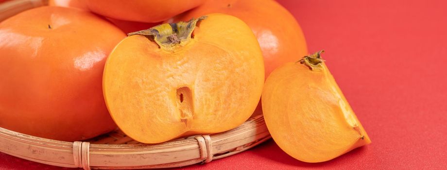 Fresh beautiful sliced sweet persimmon kaki isolated on red table background and bamboo sieve, Chinese lunar new year design concept, close up.