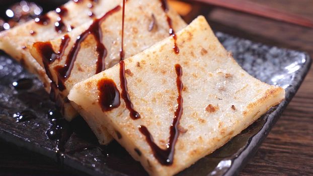 Pouring black soy sauce on ready-to-eat delicious turnip cake, Chinese traditional local dish radish cake in restaurant, close up, copy space.