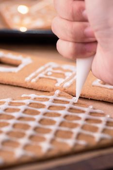 Woman is decorating gingerbread cookies house with white frosting icing cream topping on wooden table background, baking paper in kitchen, close up, macro.