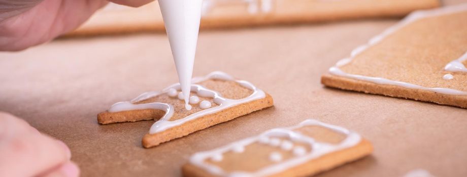 Woman is decorating gingerbread cookies house with white frosting icing cream topping on wooden table background, baking paper in kitchen, close up, macro.