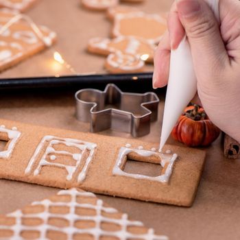 Woman is decorating gingerbread cookies house with white frosting icing cream topping on wooden table background, baking paper in kitchen, close up, macro.