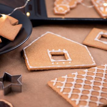 Woman is decorating gingerbread cookies house with white frosting icing cream topping on wooden table background, baking paper in kitchen, close up, macro.