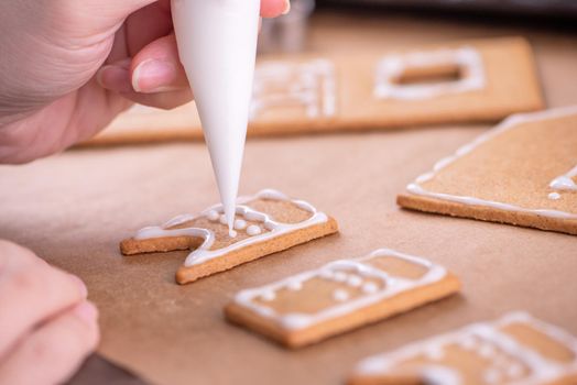 Woman is decorating gingerbread cookies house with white frosting icing cream topping on wooden table background, baking paper in kitchen, close up, macro.