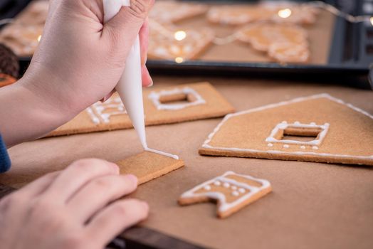 Woman is decorating gingerbread cookies house with white frosting icing cream topping on wooden table background, baking paper in kitchen, close up, macro.