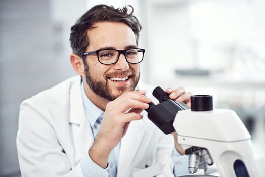 He loves every minute of his job. a cheerful young male scientist looking through a microscope while being seated inside of a laboratory