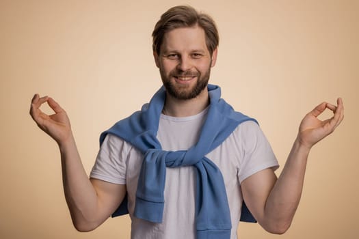 Keep calm down, relax, inner balance. Young caucasian man breathes deeply with mudra gesture, eyes closed, meditating with concentrated thoughts, peaceful mind. Guy isolated on beige background