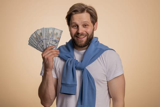 Rich pleased boss man waving money dollar cash banknotes bills like a fan, success business career, lottery winner, big income, wealth. Handsome caucasian guy isolated on beige background, indoor
