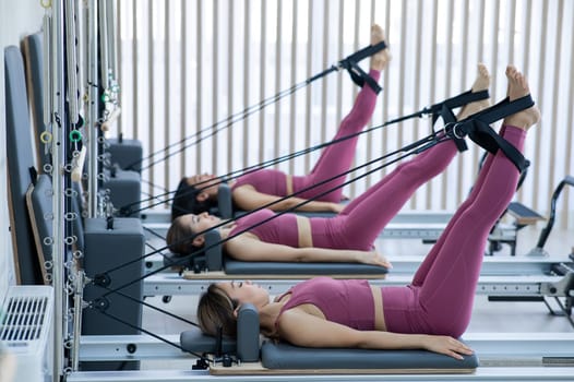 Three Asian women in pink sportswear doing pilates exercises with a reformer bed