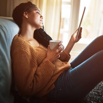 The modern way of escaping the pressures of the day. a relaxed young woman drinking coffee and using a digital tablet at home