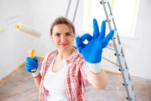 Beautiful woman holding roller painter doing ok sign with fingers while doing repair in room.