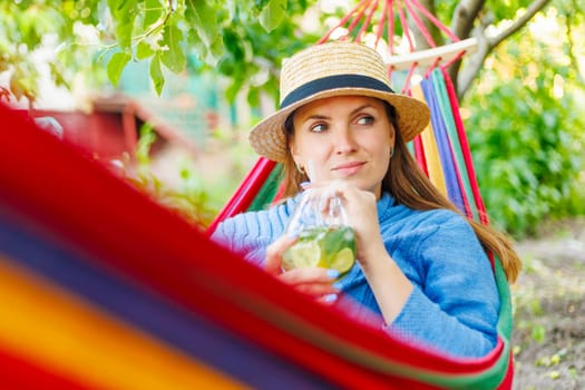 Young woman drinking a cocktail while lying in comfortable hammock at green garden.