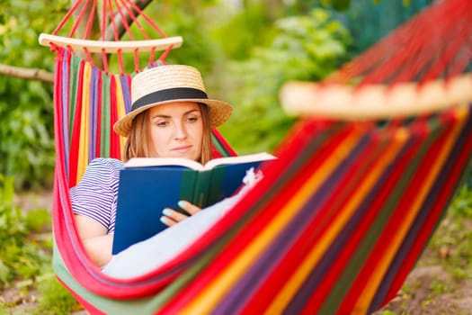 Young woman reading book while lying in comfortable hammock at green garden.