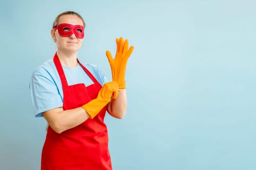 Young woman in red eye mask and apron puts on rubber gloves on blue background.