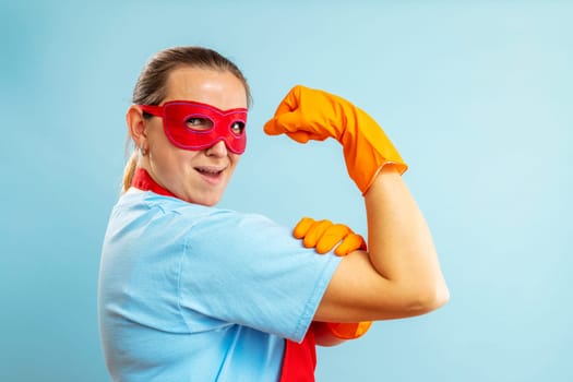 Confidence superhero house cleaner showing her powerful muscle arm with wearing red apron and eye mask on blue background