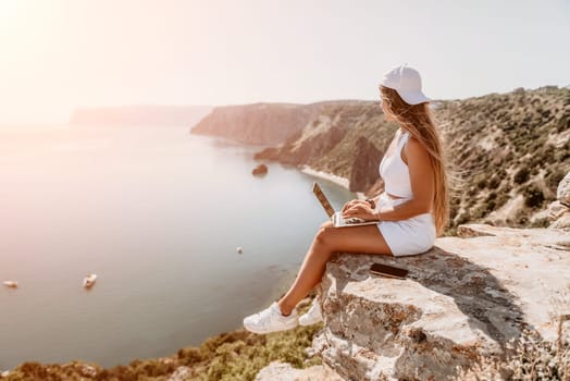 Digital nomad, Business woman working on laptop by the sea. Pretty lady typing on computer by the sea at sunset, makes a business transaction online from a distance. Freelance remote work on vacation