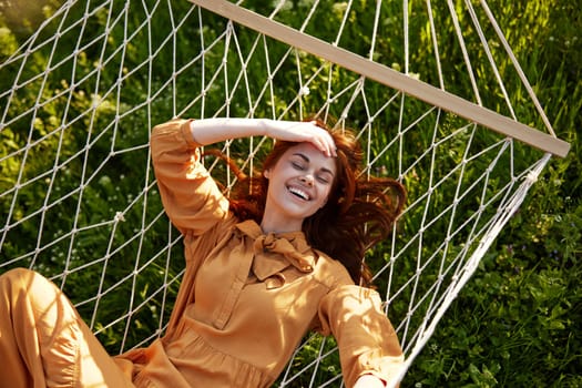 a beautiful, elegant woman lies in a long orange dress on a mesh hammock resting in nature, illuminated by the warm sunset light, holding her hand on her head, covering her face from the sun. High quality photo