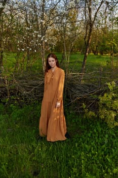 an elegant, sophisticated woman poses relaxed standing near a wicker fence at the dacha in a long orange dress, looking pleasantly at the camera. High quality photo