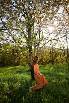 a sweet, modest, attractive woman with long red hair stands in the countryside near a flowering tree and lifts the hem of her dress up. High quality photo