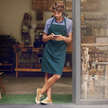 Coffee shop, typing and phone of a man as a small business owner at front door. Entrepreneur person as barista, manager or waiter in restaurant with mobile app for service, marketing or communication.