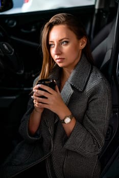 Happy woman coffee. she stands next to the car in the underground parking. Dressed in a gray coat, holding a glass of coffee in her hands, a black car