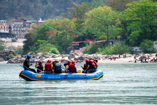 Rishikesh, Haridwar, India - circa 2023: family friends sitting in raft enjoying adventure sports while crossing in front of ghat and temple on the banks of the holy river ganga