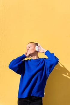 woman with a shaved head and a blue jumper enjoying happy the music in her headphones with a yellow wall in the background, concept of rhythm and positive people, copy space for text