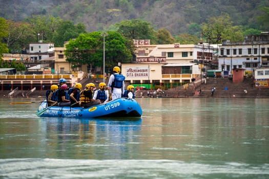 Rishikesh, Haridwar, India - circa 2023: man standing in the front of inflatable white water raft showing leadership with team in helmets and life jackets on river ganga in front of janki setu bridge with himalaya in distance