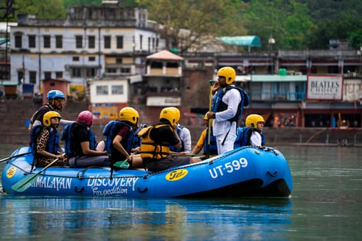 Rishikesh, Haridwar, India - circa 2023: man standing in the front of inflatable white water raft showing leadership with team in helmets and life jackets on river ganga in front of janki setu bridge with himalaya in distance