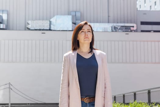 Portrait Confident Mature 40 yo Business Woman, Manager. Industrial Construction, Factory on Background. Caucasian Burnette Female With Brown Eyes Looks Away, Wears Pink Jacket, Blue Shirt.Horizontal