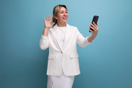 young well-groomed slender blond woman wearing a white jacket and dress uses a smartphone.