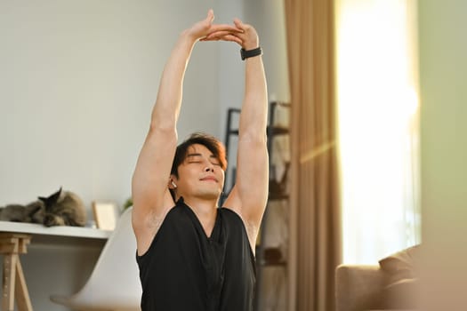 Satisfied asian man in sport outfit stretching arms resting after home workout. Healthy lifestyle and fitness concept.