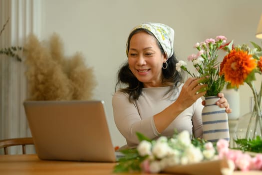 Beautiful senior woman arranging flowers and using laptop at home. Retirement, technology and lifestyle concept.